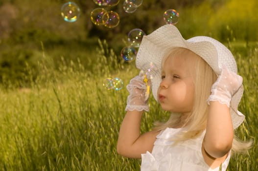 Little girl in a white dress and hat blowing soap bubbles in a field