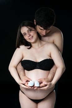 Portrait of happy young pregnant woman being hugged by husband, holding baby shoes.