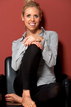 Beautiful young woman in a shirt and black trousers leaning against a red wall and sitting in a black chair