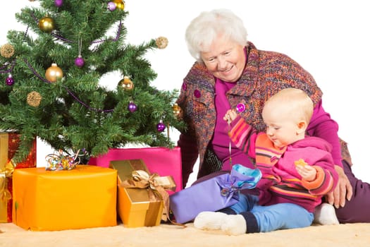 Cheerful elderly grandmother and baby sitting on the floor alongside a decorated tree unwrapping colourful Christmas gifts