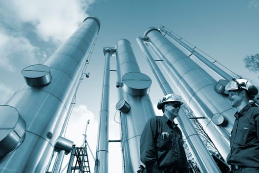 two oil and gas workers, engineers, with large gas pipelines in background, blue toning concept