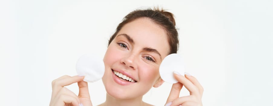 Portrait of young woman cleansing her face, takes off her makeup, washing her face, using skincare routine with cotton pads, isolated over white background.