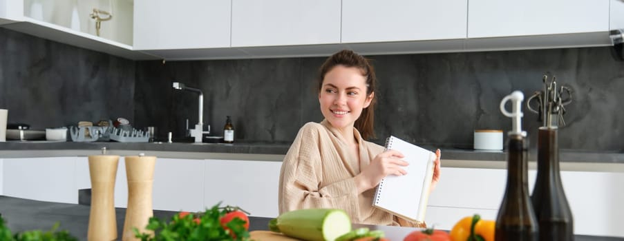 Portrait of woman writing down list of groceries, making notes in recipe, sitting in kitchen near vegetables, preparing dinner menu.