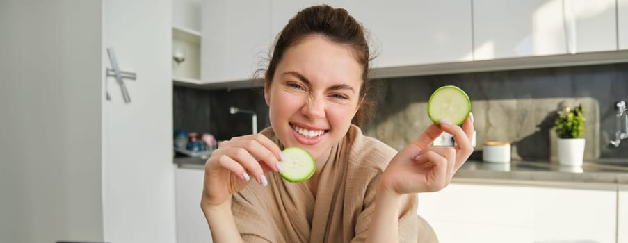 Portrait of beautiful brunette girl cooking in the kitchen, posing in bathrobe at home, holding zucchini, showing happy smile, making healthy food, vegetarian meal.