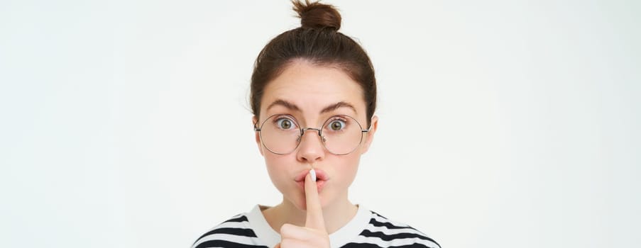 Portrait of girl with glasses, looks surprised, hughing, press finger to lips, shush shh gesture, asks to keep quiet, stands over white background.