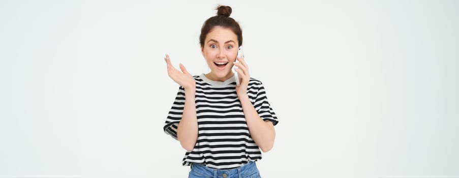 Portrait of girl answers phone call, reacts surprise, stands over white background. Communication and lifestyle concept.