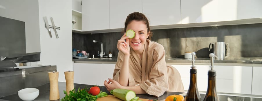 Portrait of smiling brunette woman, girl cooks vegetables, posing with peace of zucchini, leans near chopping board and looking happy while preparing meal, cooking healthy food.