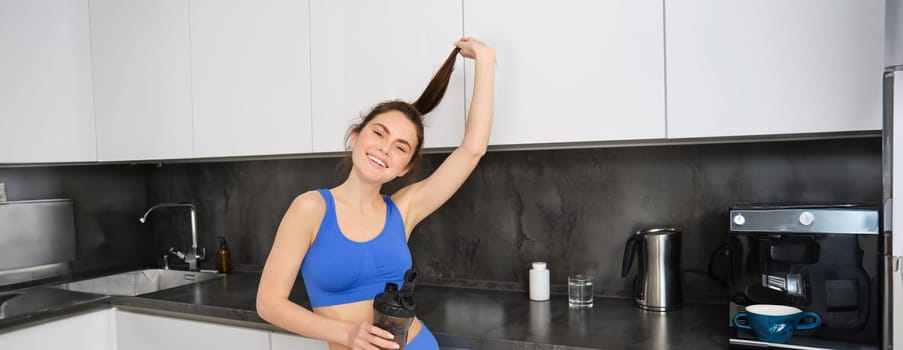 Portrait of healthy, beautiful brunette girl, wearing fitness clothing, standing in kitchen, showing her shiny hair ponytail, holding water bottle, taking dietary supplements.