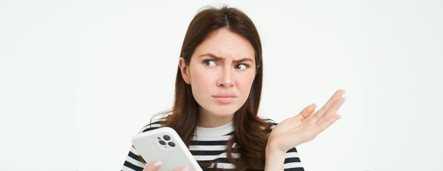 Portrait of angry, confused young woman shrugging shoulders while using mobile phone, holding smartphone with annoyed face expression, frowning, standing over white background.