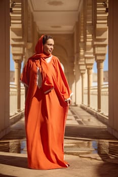 Full length portrait beautiful Middle Eastern Muslim woman in bright orange authentic dress and headscarf, looking dreamily aside, walking along the marble columns of a mosque with smoky background