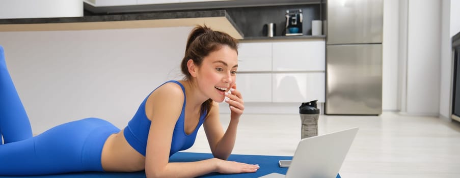 Portrait of smiling, beautiful woman sitting with laptop and watching something, doing fitness exericses from home, workout training in living room.