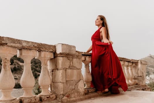 Side view a Young beautiful sensual woman in a red long dress posing on a volcanic rock high above the sea during sunset. Girl on the nature on overcast sky background. Fashion photo