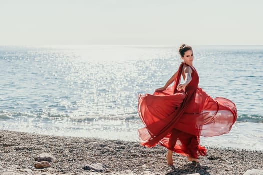 Side view a Young beautiful sensual woman in a red long dress posing on a rock high above the sea during sunrise. Girl on the nature on blue sky background. Fashion photo.