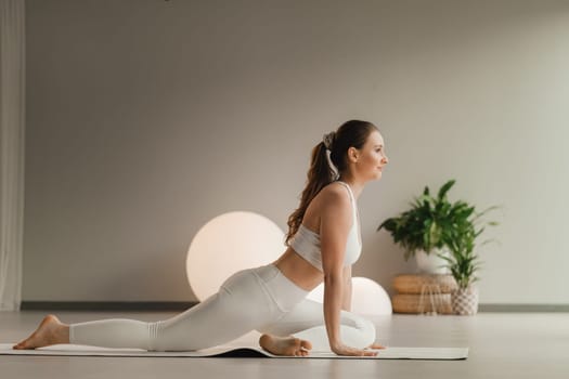 A girl in white clothes does yoga on a mat indoors.