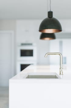 An image of a white kitchen with appliances, light fixtures and a view of the sink and faucet. The background is blurred, the focus is on the kitchen faucet. 3D rendering.