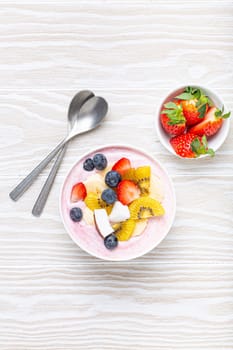 Healthy breakfast or dessert yogurt bowl with fresh banana, strawberry, blueberry, cocos, kiwi top view on rustic wooden white background with spoon