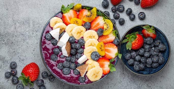 Healthy summer acai smoothie bowl with chia seeds, fresh banana, strawberry, blueberry, cocos, kiwi top view on rustic concrete background with spoon