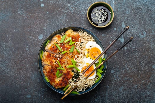 Asian noodles ramen soup with deep fried panko chicken fillet and boiled eggs in ceramic bowl with chop sticks and soy sauce on stone rustic background top view