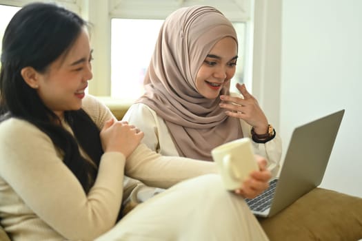 Cheerful muslim woman in hijab and friend watching funny video on laptop while sitting in cafe.