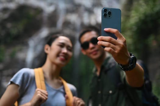 Happy couple taking selfie with smartphone against waterfall in forest. Travel and active life concept.