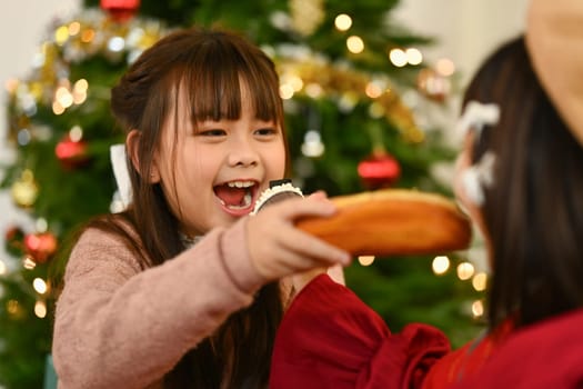 Adorable little sisters in warm winter clothes eating fresh bread near decorated Christmas tree.