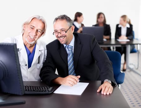 Mature male doctor showing patient prescription on his computer - Colleagues in the background