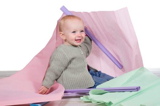 Cheerful laughing young baby with a lovely broad smile sitting on the floor amongst colourful sheets of pastel coloured paper