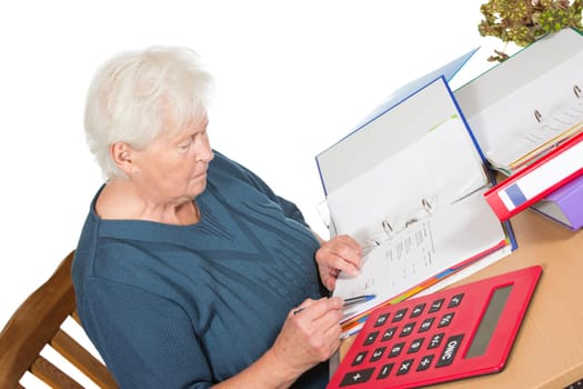 Senior woman sitting at her dining table doing her accounts checking her figures in a file with a large calculator alongside her