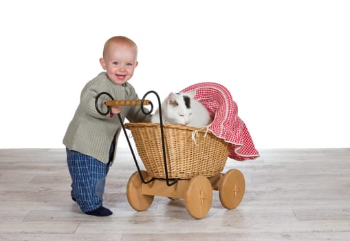 Joyful young baby standing holding the handle of a toy wicker pram with the family cat contentedly snuggled up on a blanket inside