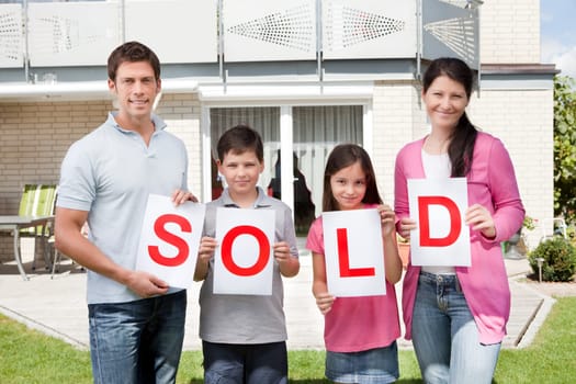 Portrait of young family holding a sold sign in front of their home