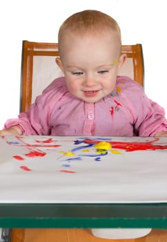 Happy baby girl sitting at a table with a colourful sheet of paint daubed paper in front of her being creative playing with paint