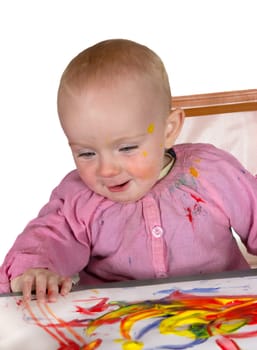 Happy baby girl daubed with colourful red and yellow paint finger painting on a large sheet of paper