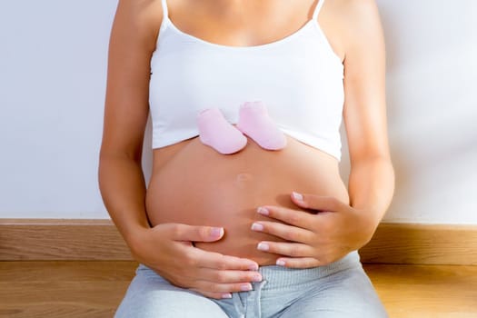Beautiful pregnant woman with baby pink shoes on her belly at home