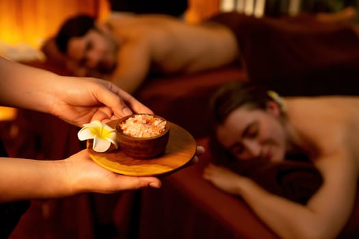 Masseur holding spa salt while couple customer having exfoliation treatment in luxury spa salon with warmth candle light ambient. Salt scrub beauty treatment in Health spa body scrub. Quiescent