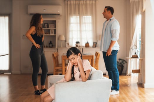 Annoyed and unhappy young girl sitting on sofa trapped in middle of tension by her parent argument in living room. Unhealthy domestic lifestyle and traumatic childhood develop to depression.Synchronos