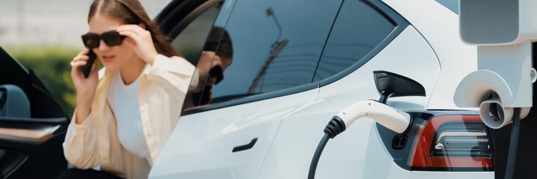 Young woman recharge EV electric vehicle at green city park while talking on phone. Sustainable urban lifestyle for environment friendly EV car with battery charging station. Panorama Expedient