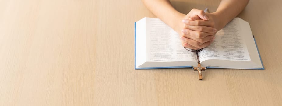 Cropped image of praying male hand holding cross on holy bible book at wooden table. Top view. Concept of hope, religion, faith, christianity and god blessing. Warm and brown background Burgeoning.