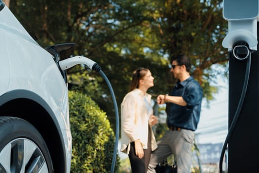 Young couple recharge electric car's battery from charging station in outdoor green city park in springtime. Rechargeable EV car for sustainable environmental friendly urban travel lifestyle.Expedient