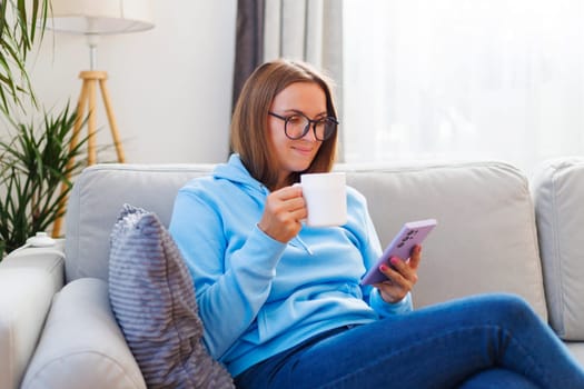 A woman in glasses relaxes on a comfortable sofa with a smartphone in hand and a cup of tea, enjoying the perfect combination of technology and tranquility.