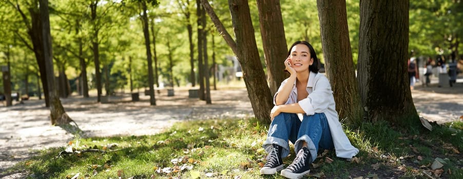 Young people. Beautiful asian girl sits near tree in park and rests, smiling and looking into distance, relaxing outdoors on fresh air in summer.