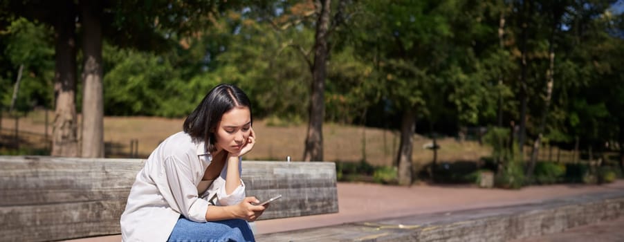 Portrait of asian girl sitting with smartphone feeling sad, looking gloomy and frustrated, waiting for a call outdoors in park.