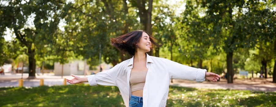 Freedom and people concept. Happy young asian woman dancing in park around trees, smiling and enjoying herself.