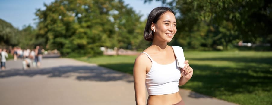 Smiling asian fitness girl holding towel on shoulder, workout in park, sweating after training exercises outdoors.