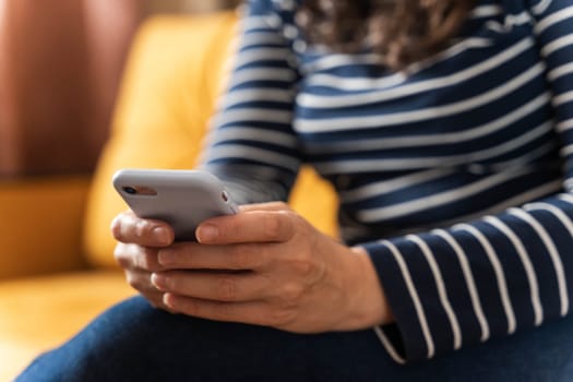 a girl in jeans and a striped blouse sits on the sofa and scrolls through pages on her smartphone screen, embodying our desire to be always up to date and socially connected.