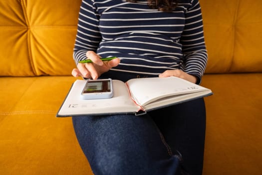 a woman makes notes in a notebook while working with a smartphone. She is sitting on the sofa in her home in a cozy atmosphere immersed in work or calculations