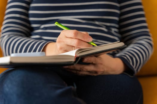 A businesswoman working in her home office and making notes in her diary. She's sitting on a yellow sofa. a woman makes notes in a notebook while working with a smartphone