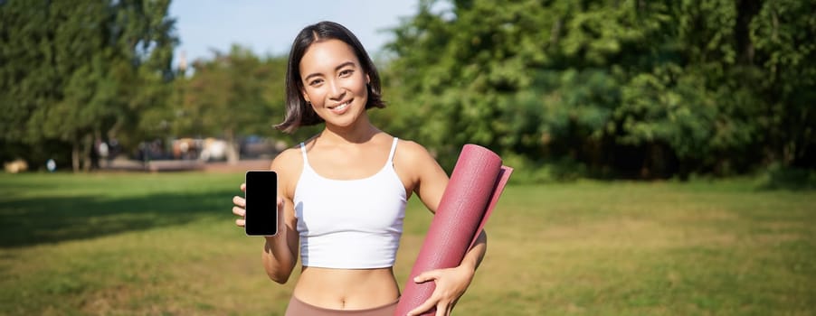 Smiling asian fitness girl with rubber yoga mat, shows her smartphone screen, recommends workout application, stands on lawn in park.