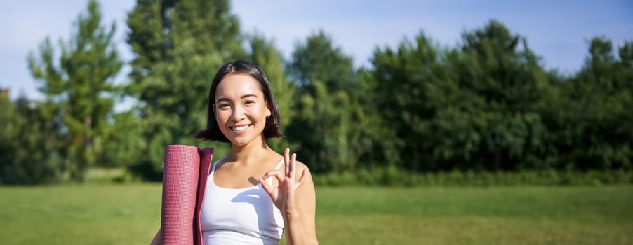 Excited young woman standing with sports mat, yoga clothes, shows okay sign, workout in park, wellbeing training session outdoors.