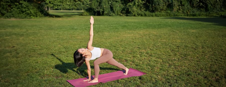 Portrait of young asian woman stretching, doing yoga on rubber mat, exercising in park, mindful training on fresh air.