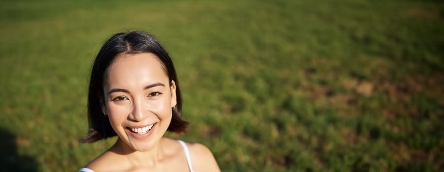 Vertical shot of young asian woman doing yoga, practice mindfulness, smiling and looking relaxed, sitting on rubber mat in park.